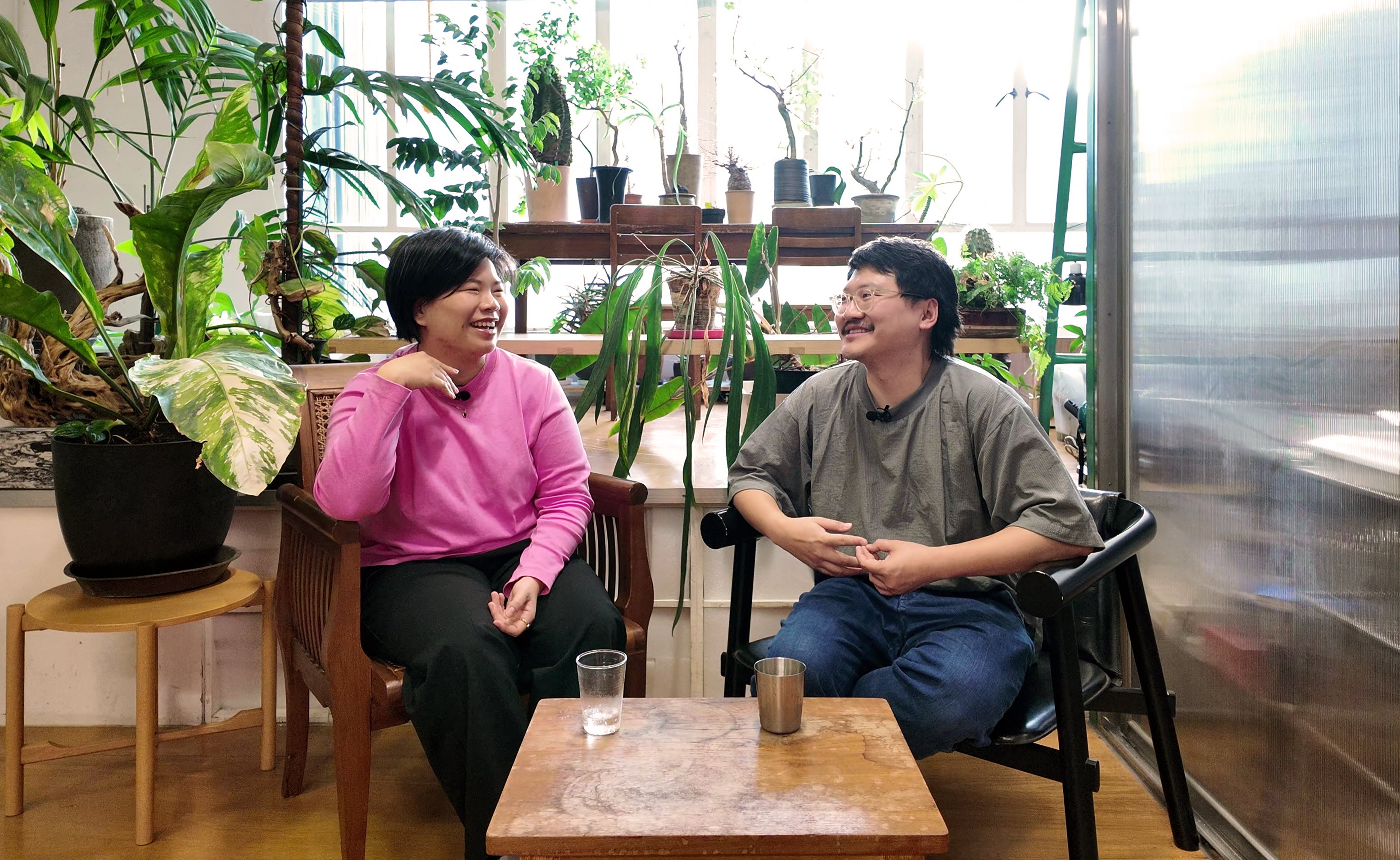 In a room with many green plants, there is a woman in a pink sweater on the left seated next to a man in a green shirt. They are laughing.